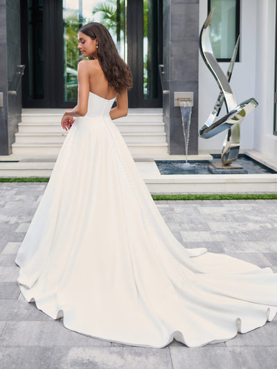 A woman in a Simple Satin Ball Gown with Pearl Bow Bodice Detail Wedding Dress by Adrianna Papell Platinum, offered by Bergamot Bridal, stands in front of a modern building with a staircase and metal sculpture.