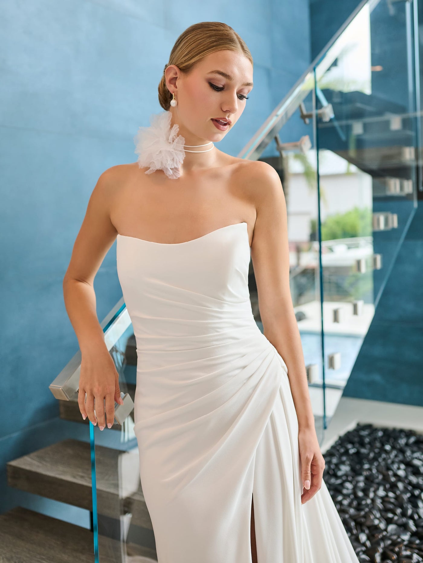 A person wearing the Draped Elegant Crepe Fit & Flare Wedding Gown with Lace Up Back by Adrianna Papell Platinum from Bergamot Bridal, featuring a chic slit and strapless design, stands by a modern staircase. They have short, elegantly styled hair and are adorned with large floral earrings.