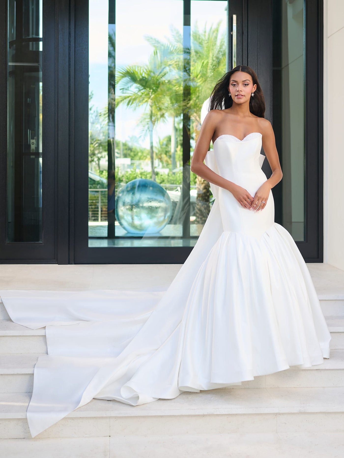 A woman wearing the Elegant Mermaid Wedding Dress with Oversized Bow Detail and Full Skirt by Adrianna Papell Platinum from Bergamot Bridal poses on steps in front of a glass door, with palm trees visible in the background.