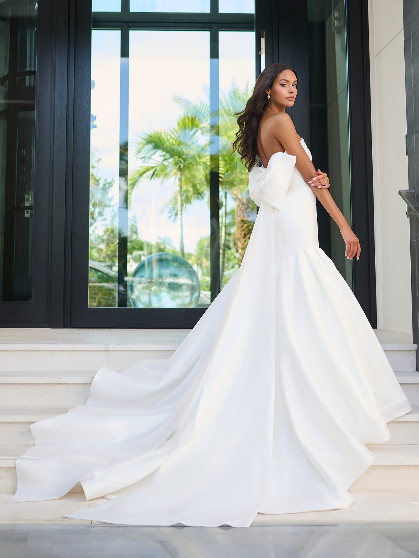 A woman in a breathtaking Elegant Mermaid Wedding Dress by Adrianna Papell Platinum, featuring an oversized bow detail and full skirt from Bergamot Bridal, stands gracefully on steps near large glass doors, with lush greenery visible in the background.