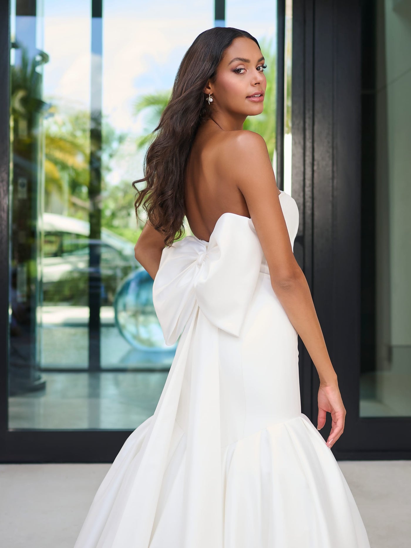 A woman in the Elegant Mermaid Wedding Dress with Oversized Bow Detail and Full Skirt by Adrianna Papell Platinum from Bergamot Bridal stands in front of a modern glass doorway, showcasing its strapless style and pearl and crystal encrusted detachable bow.