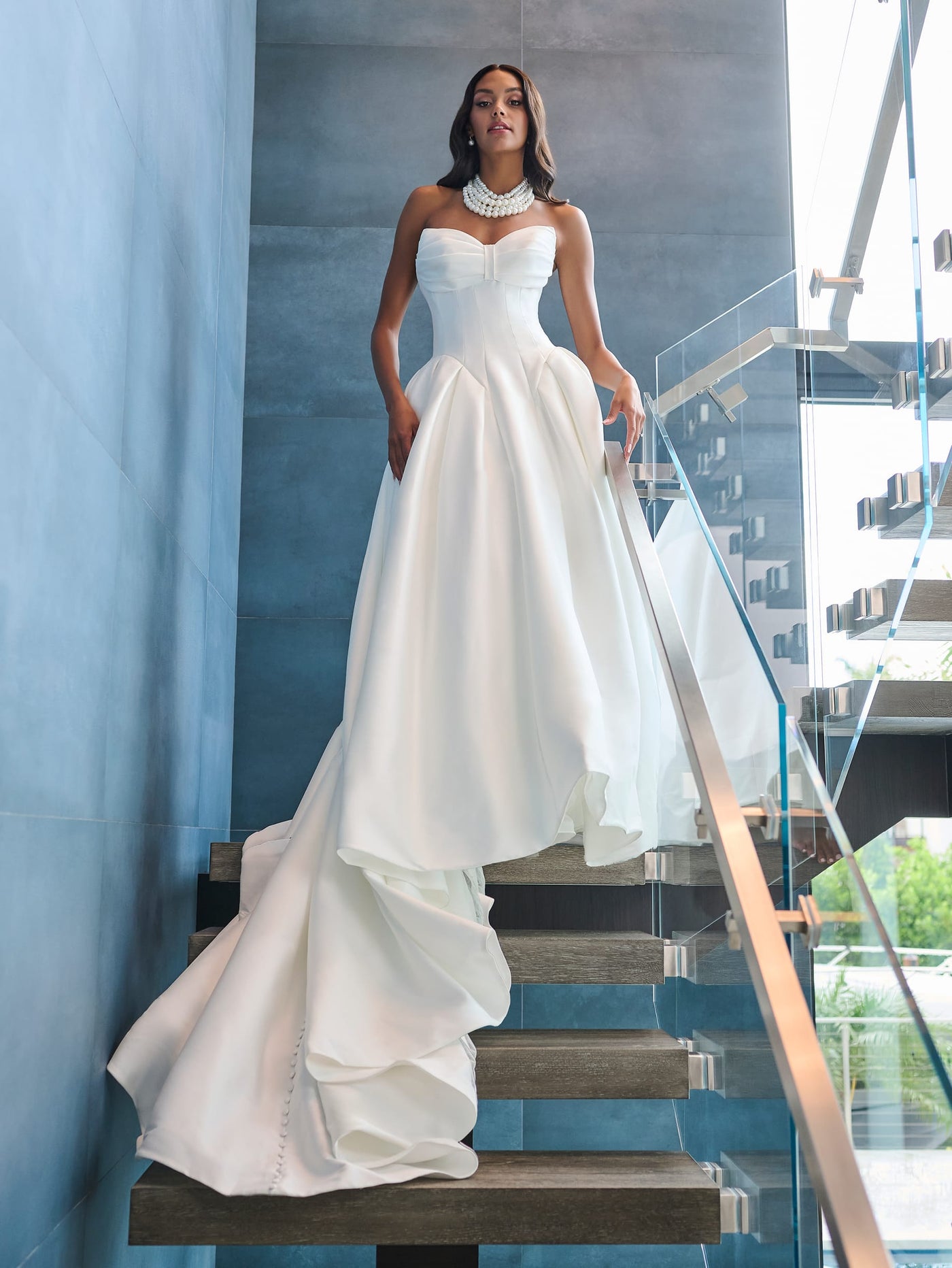 Draped in the elegant Drop Waist Princess Ballgown with Unique Neckline Wedding Dress by Adrianna Papell Platinum from Bergamot Bridal, a woman stands gracefully on a modern staircase.