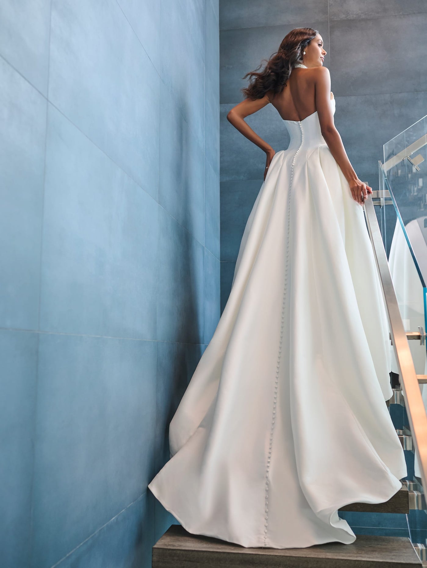 Wearing the Drop Waist Princess Ballgown with Unique Neckline Wedding Dress by Adrianna Papell Platinum from Bergamot Bridal, a woman gracefully descends a modern staircase framed by blue-gray walls.