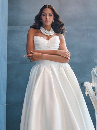 A woman stands in a Drop Waist Princess Ballgown with a unique neckline by Adrianna Papell Platinum, available at Bergamot Bridal. The strapless white wedding dress features a full, pleated skirt and is paired with a chunky pearl necklace. She poses against a gray textured wall with crossed arms and gazes forward.