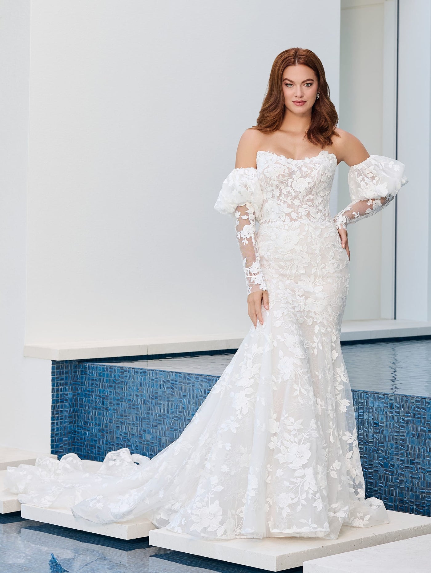 A woman wearing the Floral Lace Mermaid Wedding Dress with Scoop Neckline and Illusion Back by Adrianna Papell Platinum from Bergamot Bridal stands near a tiled water feature, the scoop neckline highlighting her timeless elegance.