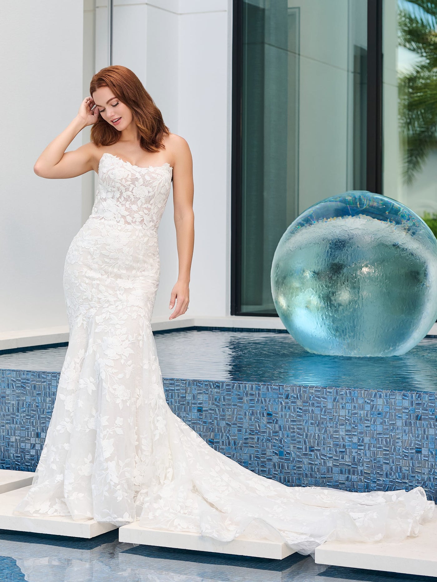 A woman in a Floral Lace Mermaid Wedding Dress with a scoop neckline and illusion back by Adrianna Papell Platinum from Bergamot Bridal poses gracefully beside a blue-tiled pool, holding a large glass orb.