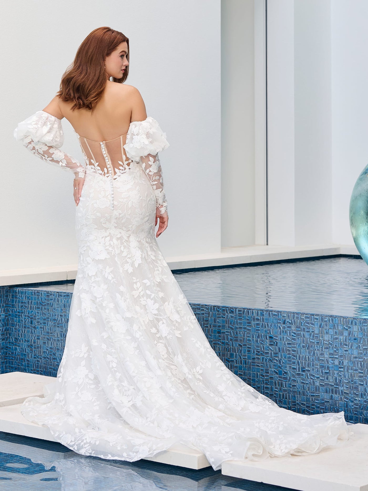 A woman in a Floral Lace Mermaid Wedding Dress with a scoop neckline and illusion back by Adrianna Papell Platinum from Bergamot Bridal, adorned with puff sleeves and a long train, stands gracefully by the pool.