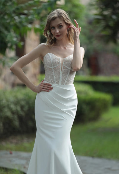 A woman in a Bergamot Bridal Plunging Sweetheart Neckline Beaded Crepe Fit And Flare wedding dress posing in a garden, with her hand gracefully touching her hair.