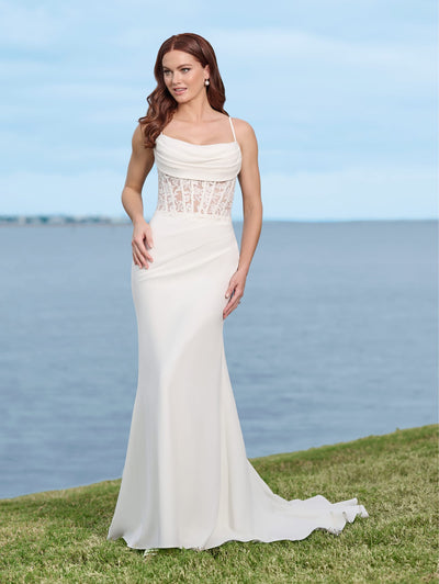 A woman in the Sexy Stretch Crepe Fit & Flare Bridal Gown with Sheer Lace Bodice and Cowl Neckline by Adrianna Papell Platinum from Bergamot Bridal stands on grass near a body of water, looking to the side.