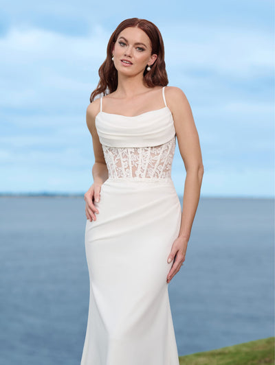 A woman in a white Sexy Stretch Crepe Fit & Flare Bridal Gown with Sheer Lace Bodice and Cowl Neckline by Adrianna Papell Platinum from Bergamot Bridal stands outdoors near a body of water, with the blue sky providing a stunning backdrop.