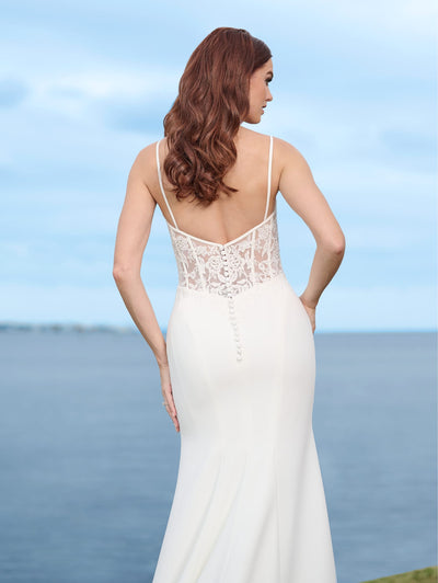 A woman in a Sexy Stretch Crepe Fit & Flare Bridal Gown with Sheer Lace Bodice and Cowl Neckline by Adrianna Papell Platinum, from Bergamot Bridal, stands with her back to the camera, facing a large body of water.