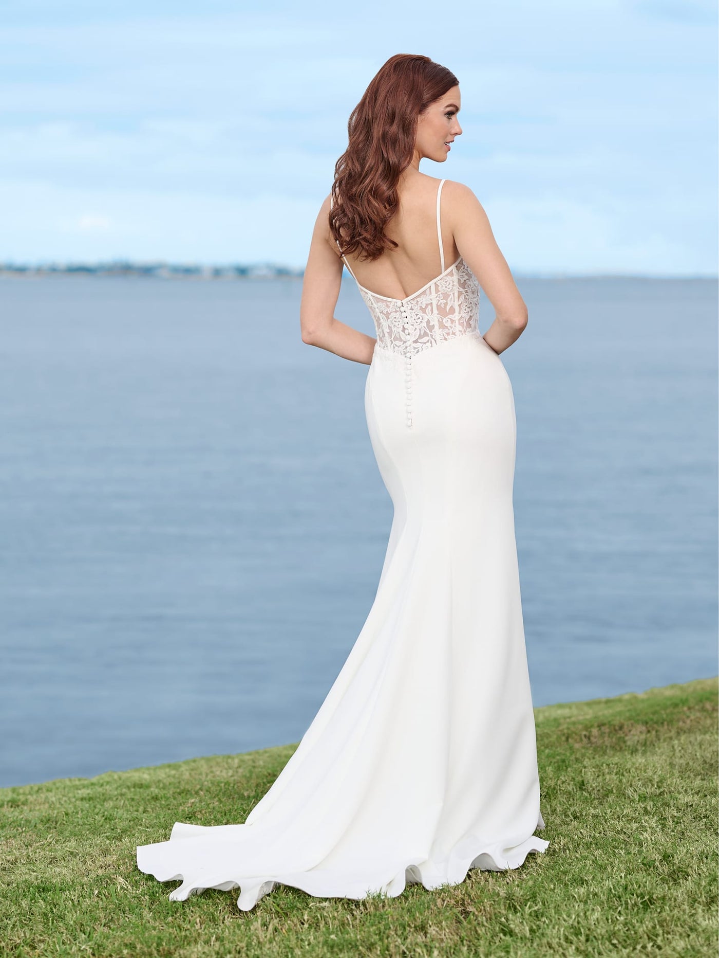 A woman wearing the Sexy Stretch Crepe Fit & Flare Bridal Gown with Sheer Lace Bodice and Cowl Neckline by Adrianna Papell Platinum from Bergamot Bridal stands on grass by a body of water, facing away.