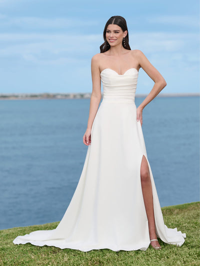 A woman in the Simple A-line Wedding Dress with a Beaded Sweetheart Neckline and Thigh Slit by Adrianna Papell Platinum from Bergamot Bridal stands on grass, framed by the blue sea and sky backdrop.