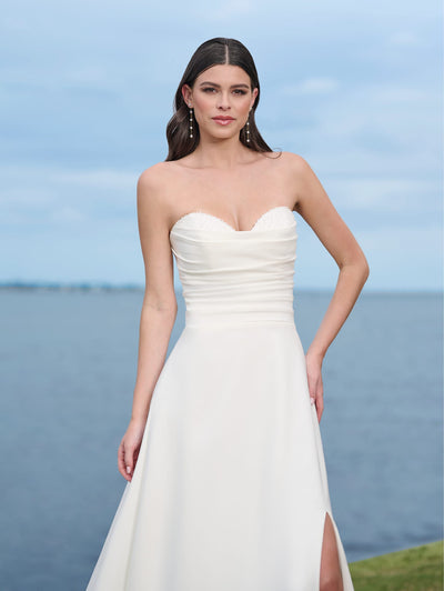 A woman in a Simple A-line Wedding Dress with a beaded sweetheart neckline and thigh slit by Adrianna Papell Platinum for Bergamot Bridal poses outdoors by the water on a cloudy day.