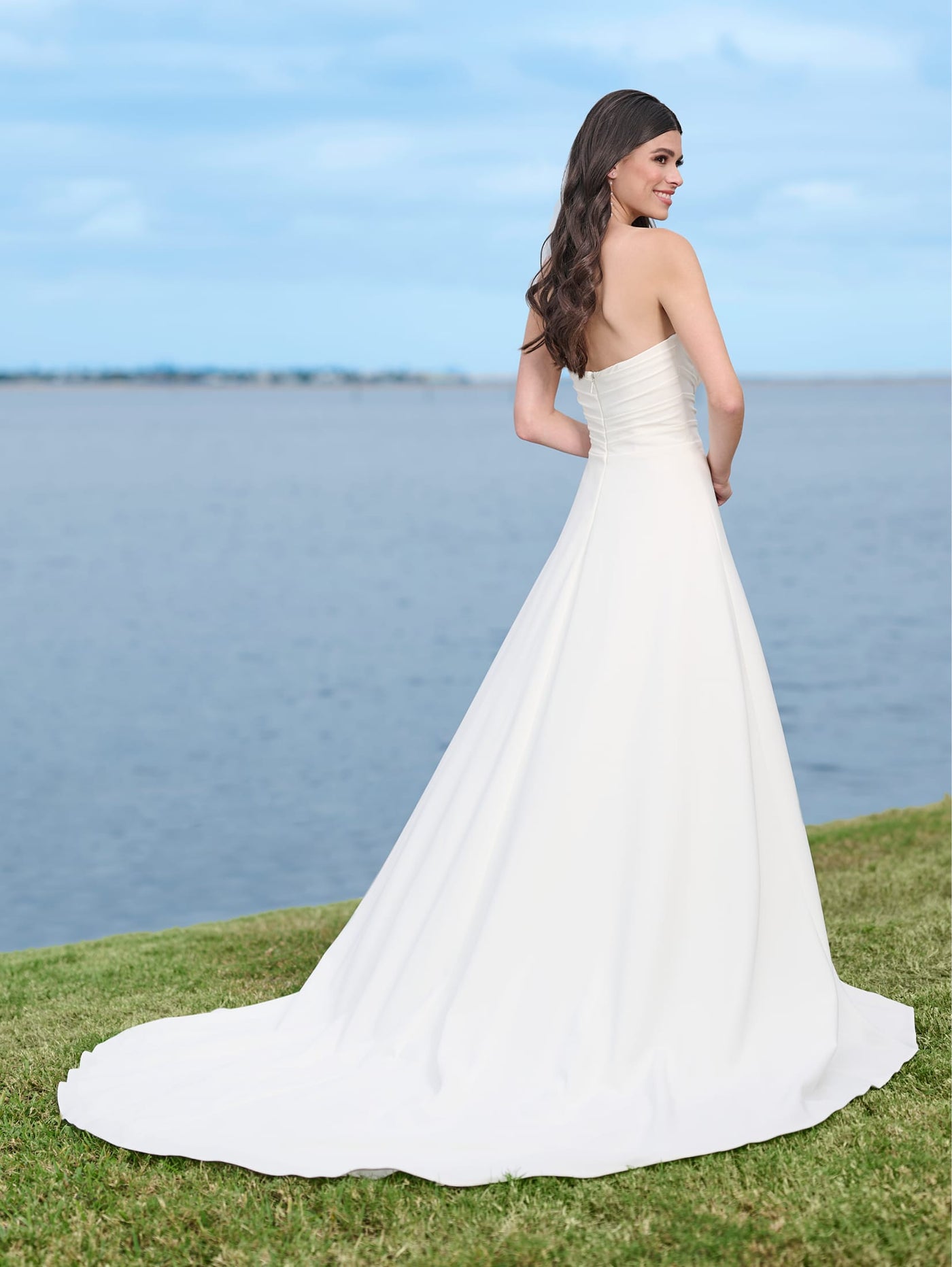 Wearing a Simple A-line Wedding Dress with a Beaded Sweetheart Neckline and Thigh Slit by Adrianna Papell Platinum, a woman from Bergamot Bridal stands on grass near a body of water with her back facing the camera.