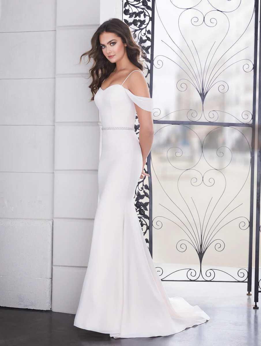 A woman in an elegant white off-the-shoulder wedding dress with a beaded belt stands near a decorative iron gate wearing the Bergamot Bridal Paloma Blanca 4854 Simple Fit and Flare Gown - Off The Rack.