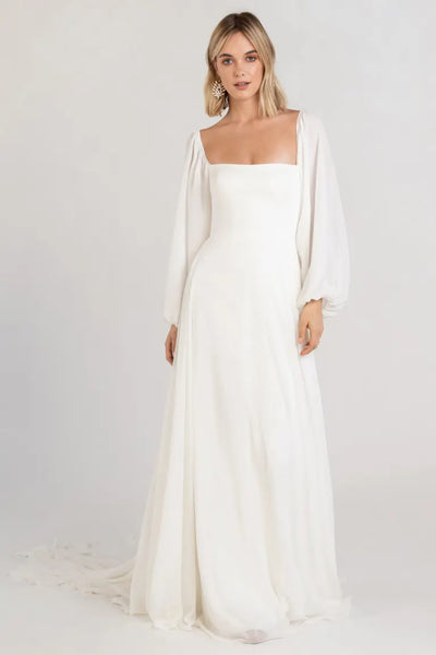 Woman in a long-sleeved Louise - Jenny Yoo wedding dress by Bergamot Bridal standing against a neutral background.