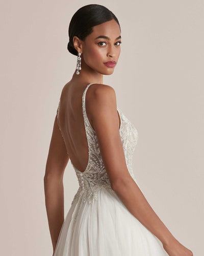 A woman in an elegant Justin Alexander Cady Dress - Off The Rack from Bergamot Bridal with a beaded bodice and open back looks over her shoulder, wearing dangling earrings.