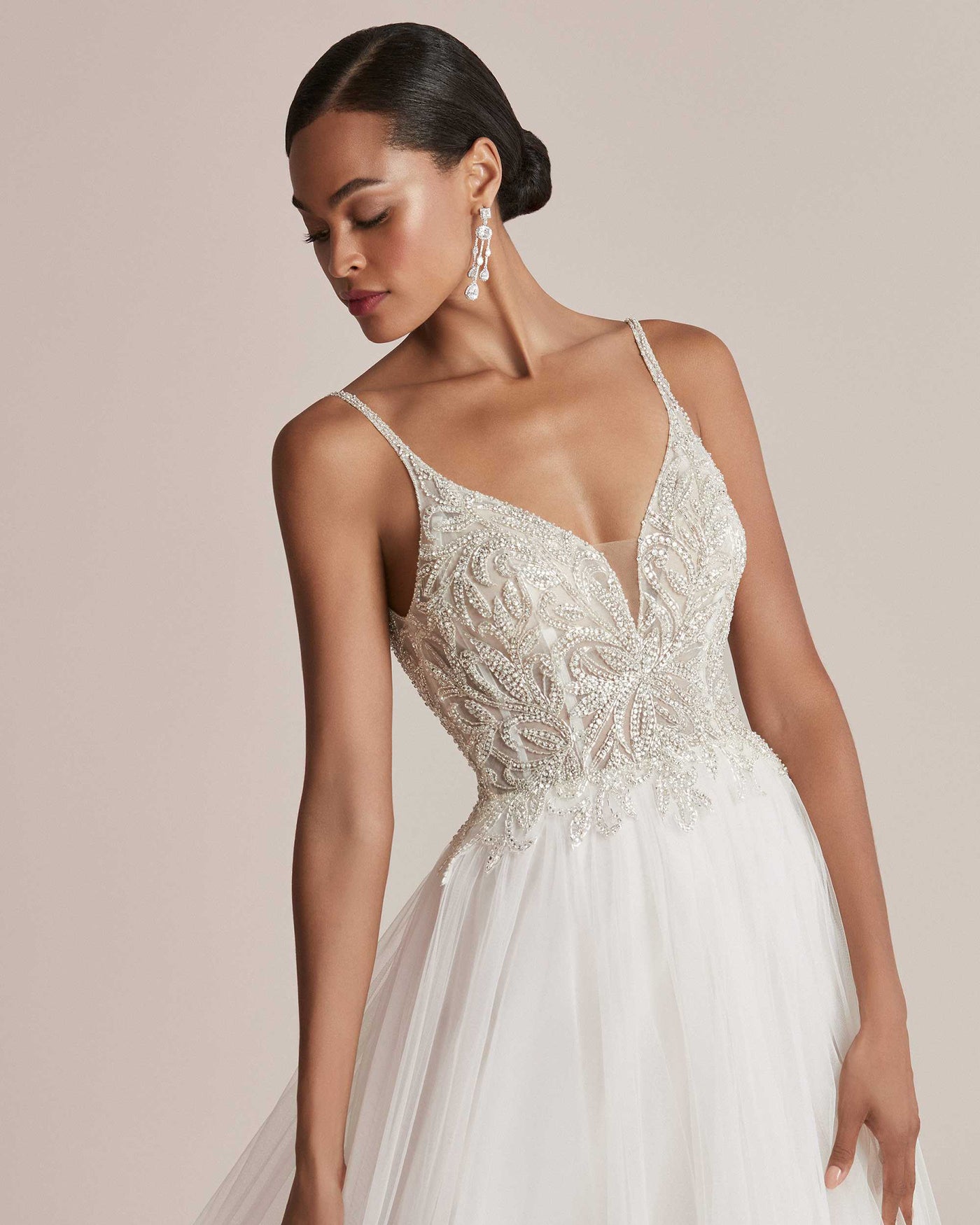 A woman models an elegant Justin Alexander Cady Dress - Off The Rack from Bergamot Bridal with intricate beading on the bodice and a flowing tulle skirt. She wears sparkling earrings.