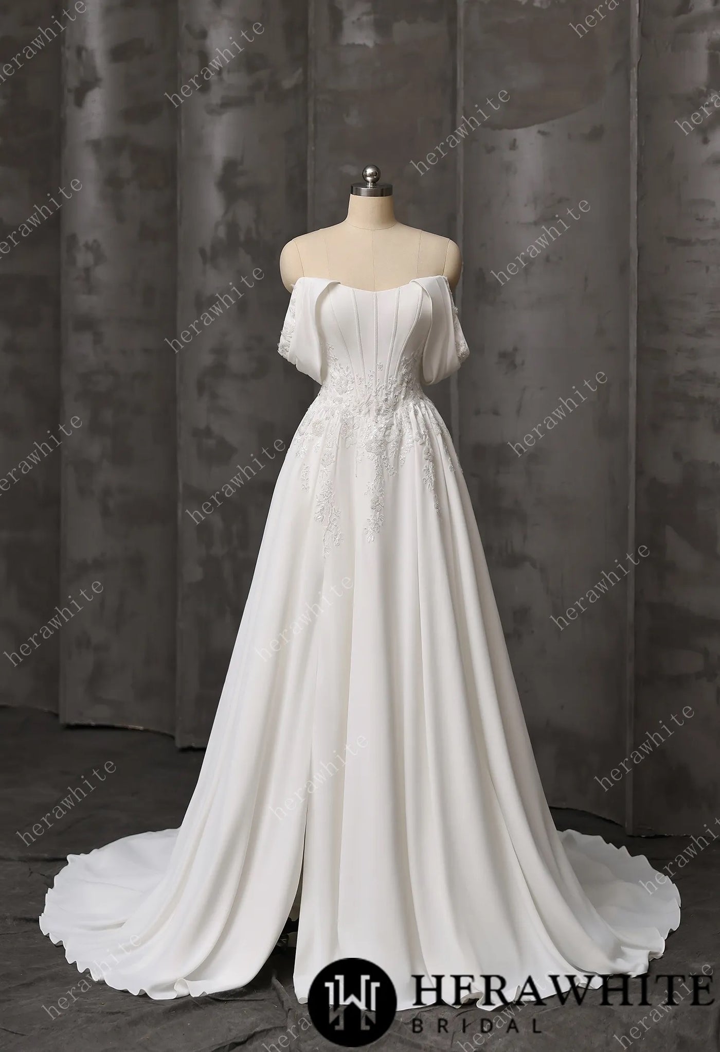 The Coloured Embroidered A-Line Gown by Bergamot Bridal features a fitted bodice, floral lace details, cat eye scoop neckline, and a flowing train, displayed on a dress form against a gray backdrop.