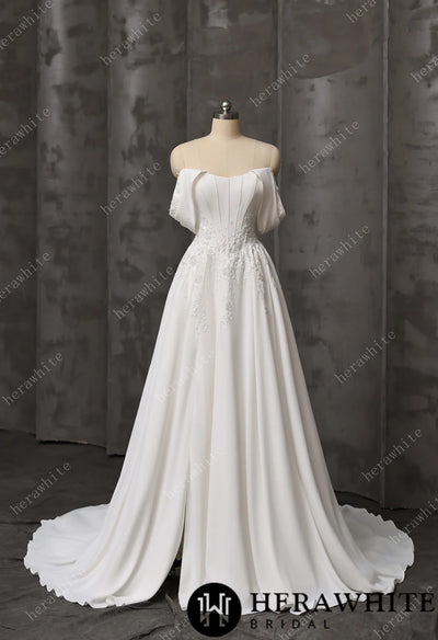 The Coloured Embroidered A-Line Gown by Bergamot Bridal features a fitted bodice, floral lace details, cat eye scoop neckline, and a flowing train, displayed on a dress form against a gray backdrop.