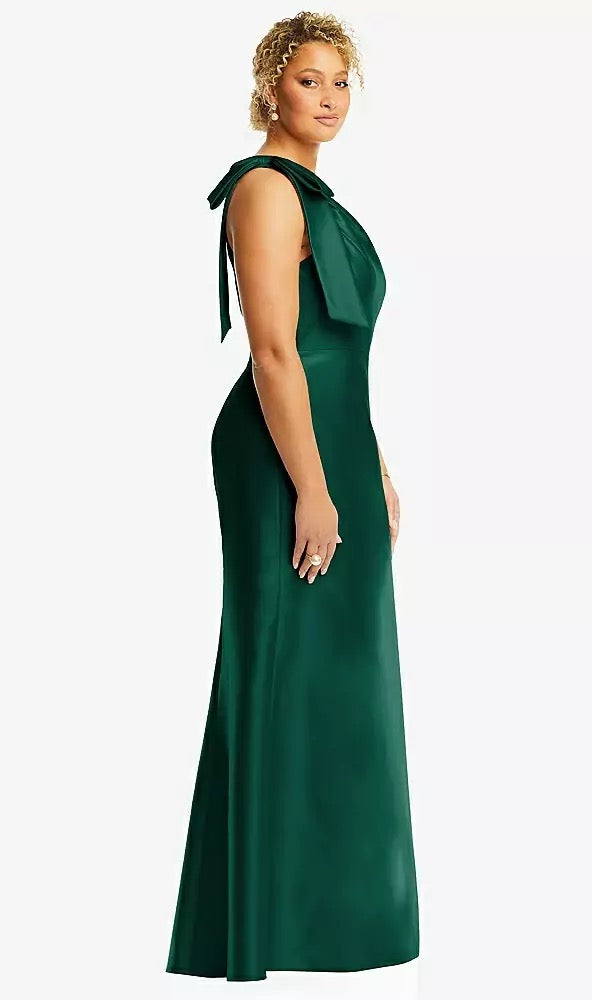 A woman stands against a white background wearing the Bow One-Shoulder Satin Trumpet Gown Bridesmaid Dress in dark green from Bergamot Bridal's Dessy collection, featuring a satin twill finish and an elegant bow on the shoulder.