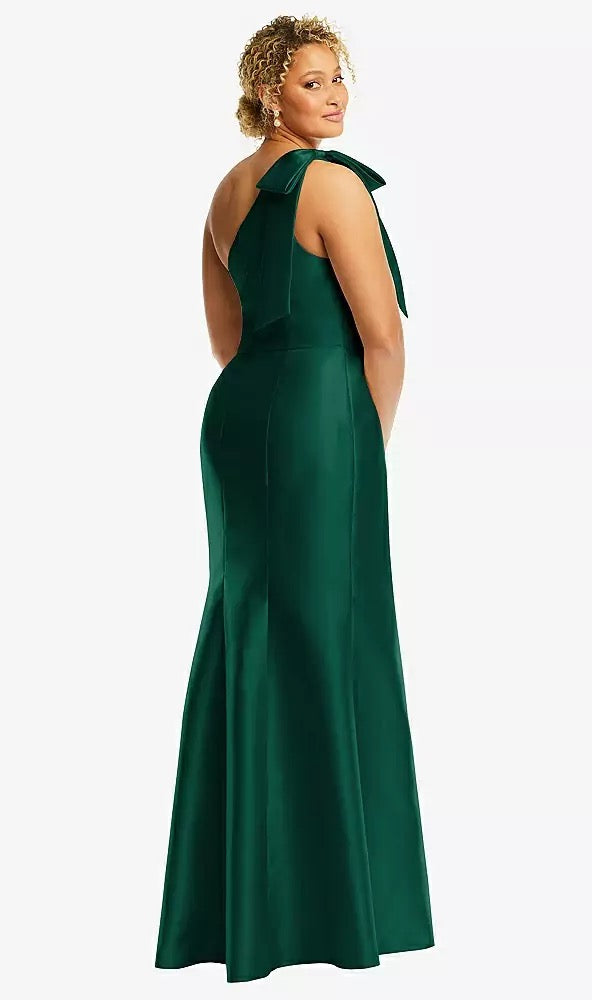 A woman poses partially away from the camera wearing a Bergamot Bridal's Bow One-Shoulder Satin Trumpet Gown Bridesmaid Dress, Style D794 by Dessy, in green satin twill with a bow detail on the single shoulder.