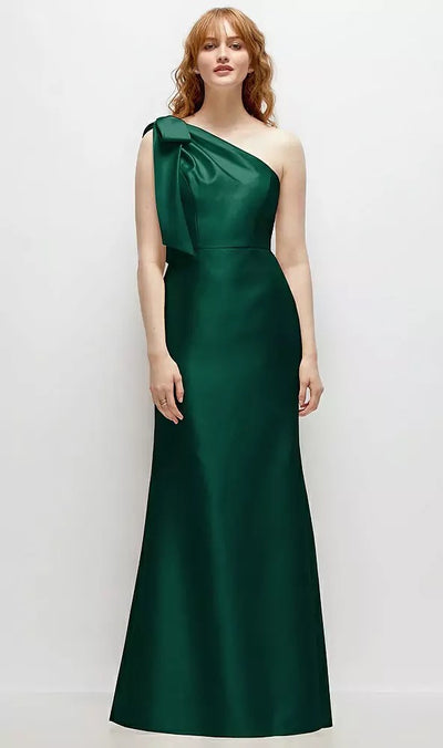 A person wearing the Bow One-Shoulder Satin Trumpet Gown Bridesmaid Dress - Style D794 by Dessy from Bergamot Bridal, showcasing its elegant dark green satin twill and distinctive bow detail, stands against a plain white background.