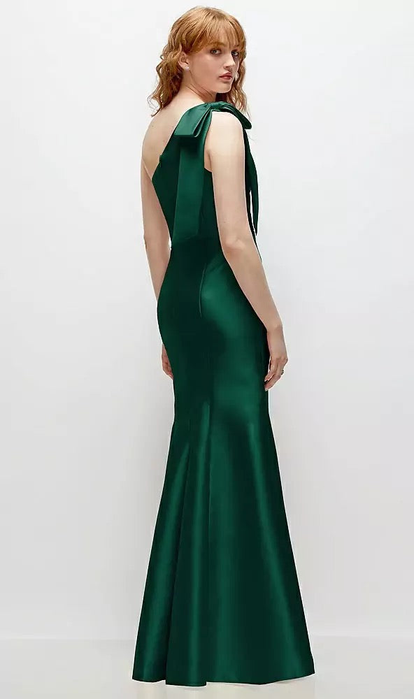 A person in the Bow One-Shoulder Satin Trumpet Gown Bridesmaid Dress by Bergamot Bridal, featuring a backless design and crafted from dark green satin twill, poses against a plain white background.