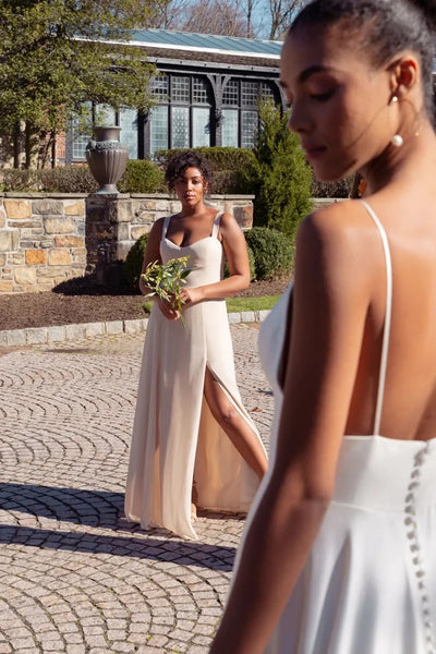 A woman in a Harris chiffon bridesmaid dress by Jenny Yoo from Bergamot Bridal holding a small bouquet stands in the background, while another woman with her back to the camera, wearing a similar dress with a bombshell neckline, is