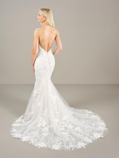 A person elegantly wears the Delicate Lace Fit & Flare Wedding Dress by Adrianna Papell Platinum from Bergamot Bridal, featuring a backless design with a floral lace mermaid silhouette, a sweetheart neckline, and delicate lace appliqué. A sheer petal train gracefully extends behind them as they face away.