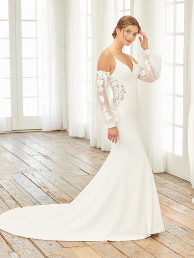 A woman gracefully stands on a wooden floor in front of tall, elegant windows, wearing the Crepe Fit & Flare Bridal Gown with Lace Back and Side Cut-Outs by Adrianna Papell Platinum from Bergamot Bridal.