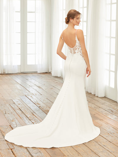 A woman wearing the Crepe Fit & Flare Bridal Gown with Lace Back and Side Cut-Outs by Adrianna Papell Platinum from Bergamot Bridal poses elegantly on a wooden floor, surrounded by large windows adorned with white curtains. This stunning sleeveless wedding dress features a plunging neckline and a long train, combining crepe and lace for an exquisite fit n' flare silhouette.