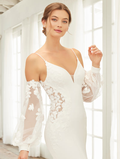 A woman gracefully stands in front of a window wearing the stunning Crepe Fit & Flare Bridal Gown with Lace Back and Side Cut-Outs by Adrianna Papell Platinum from Bergamot Bridal. This gown features a fit n' flare silhouette and exquisite floral lace details, while sheer off-the-shoulder sleeves perfectly complement the plunging neckline, creating an elegant and timeless look.