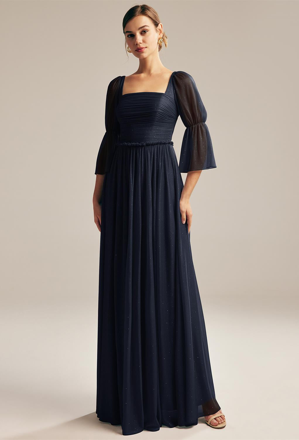 A woman in a Liza - Glitter Mesh Bridesmaid Dress by Bergamot Bridal, with sheer puffed sleeves, suitable for wedding dresses, standing against a neutral background.