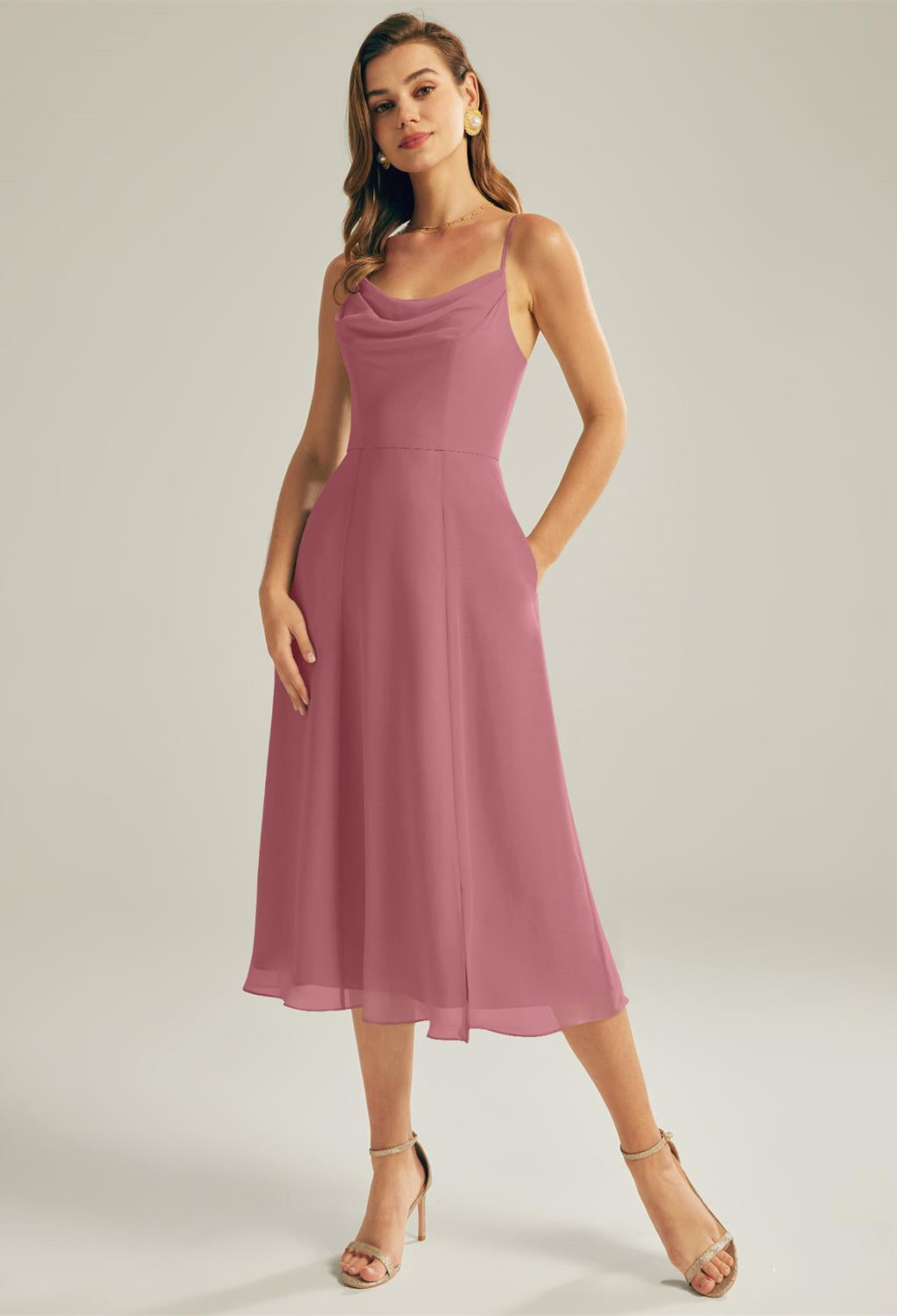 Against a neutral backdrop, a woman poses in strappy heels and the "Maribel - Chiffon Bridesmaid Dress - Off the Rack" by Bergamot Bridal, which showcases its pink, sleeveless midi design that seems to have just emerged from a bridal shop in London, Ontario. Her stunning outfit would elegantly complement affordable wedding or bridesmaid dresses.