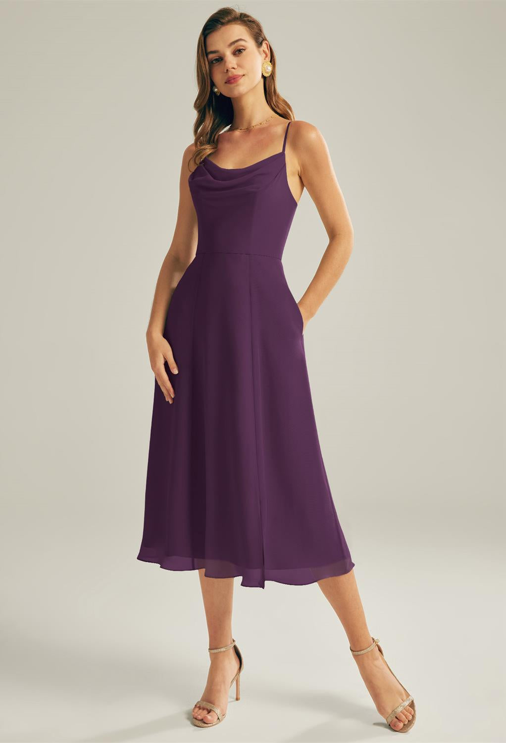 A woman wearing the sleeveless Maribel chiffon bridesmaid dress by Bergamot Bridal, in a striking purple hue, stands gracefully with her hands in the pockets against a neutral background, capturing the essence of chic off-the-rack bridal fashion from a leading shop in London, Ontario.