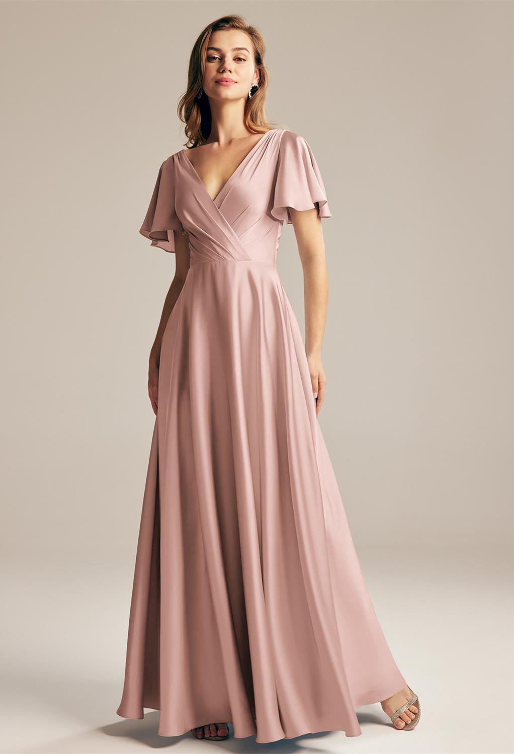 A woman in a long, flowing pink dress from Bergamot Bridal stands against a plain background, embodying the elegance found in the Furst - Satin Charmeuse Bridesmaid Dress - Off The Rack selection from an affordable bridal shop in London, Ontario. Looking forward, she captures the essence of timeless bridesmaid dresses.