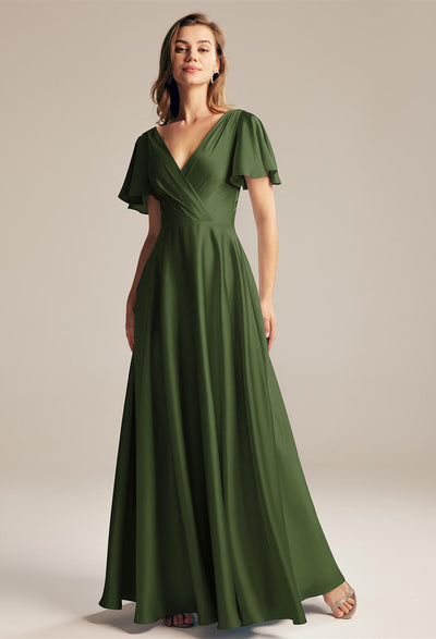 A woman in a flowing green dress stands confidently against a neutral background, reminiscent of the elegant Furst Satin Charmeuse Bridesmaid Dress by Bergamot Bridal, available off the rack at bridal shops in London, Ontario.