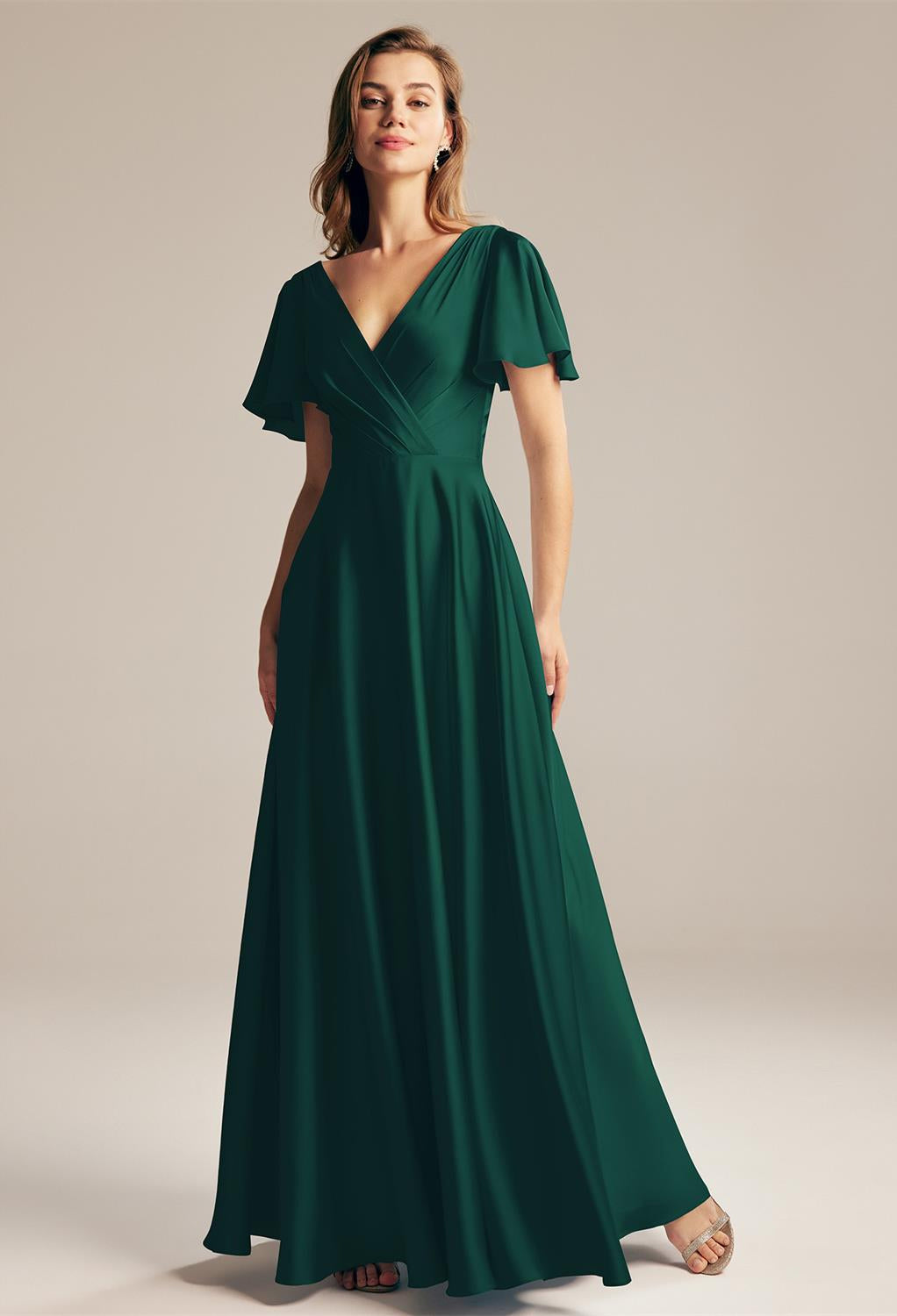 A person stands elegantly in a floor-length, dark green dress with short, flowy sleeves, similar to the Furst - Satin Charmeuse Bridesmaid Dress by Bergamot Bridal, reminiscent of those found in a chic bridal shop in London, Ontario.