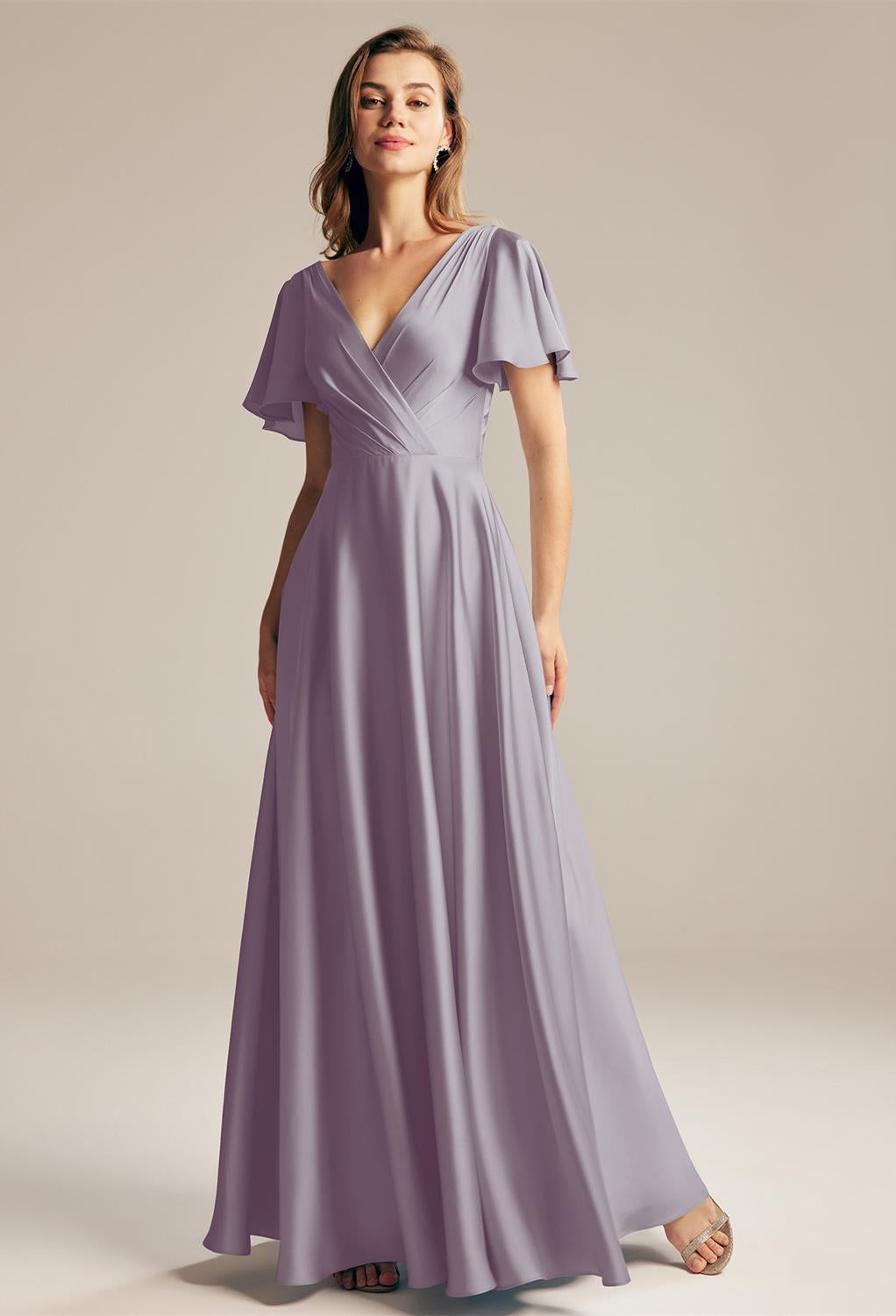 A woman in a long, flowing lavender dress stands smiling against a neutral background, reminiscent of the elegant Furst Satin Charmeuse Bridesmaid Dress by Bergamot Bridal, available off the rack at the bridal shop in London, Ontario.