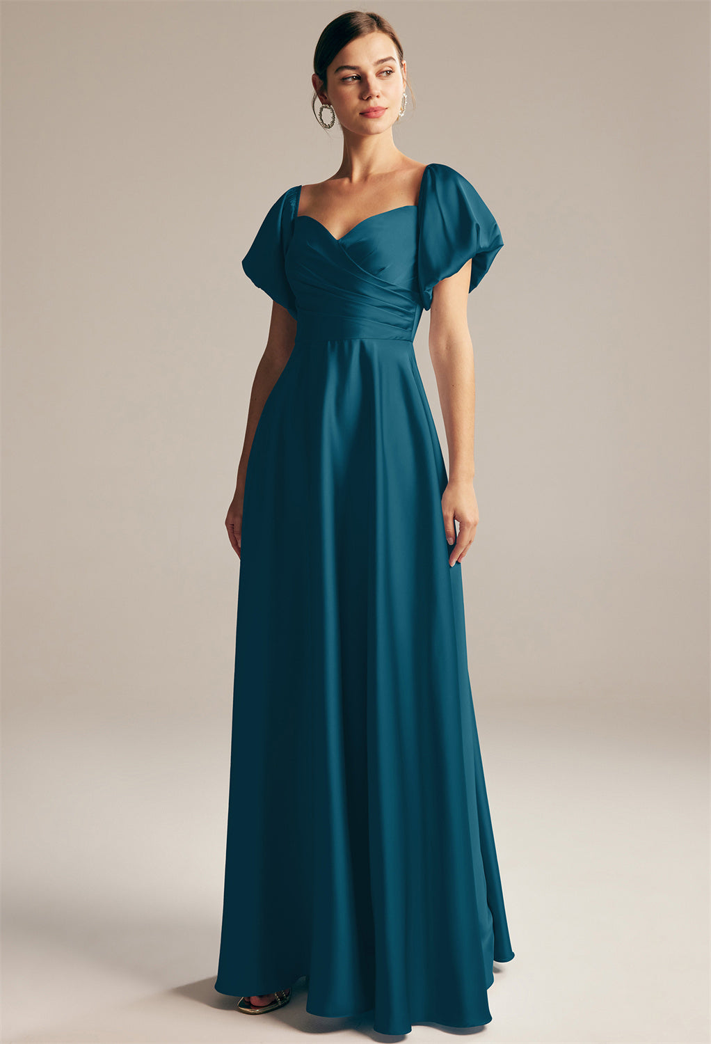 A person in a satin charmeuse bridesmaid dress by Bergamot Bridal, known as the "Dey," stands gracefully against a neutral background, evoking the elegant styles found in bridal shops in London, Ontario. The dress features a stunning teal color and puff sleeves.