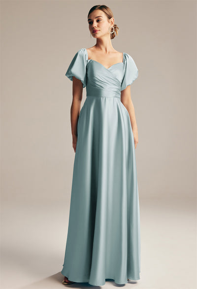 A woman wearing the Dey - Satin Charmeuse Bridesmaid Dress by Bergamot Bridal stands in front of a plain background, displaying elegant bridal shop styles from London, Ontario. The dress is light blue, floor-length, and features short puffed sleeves.