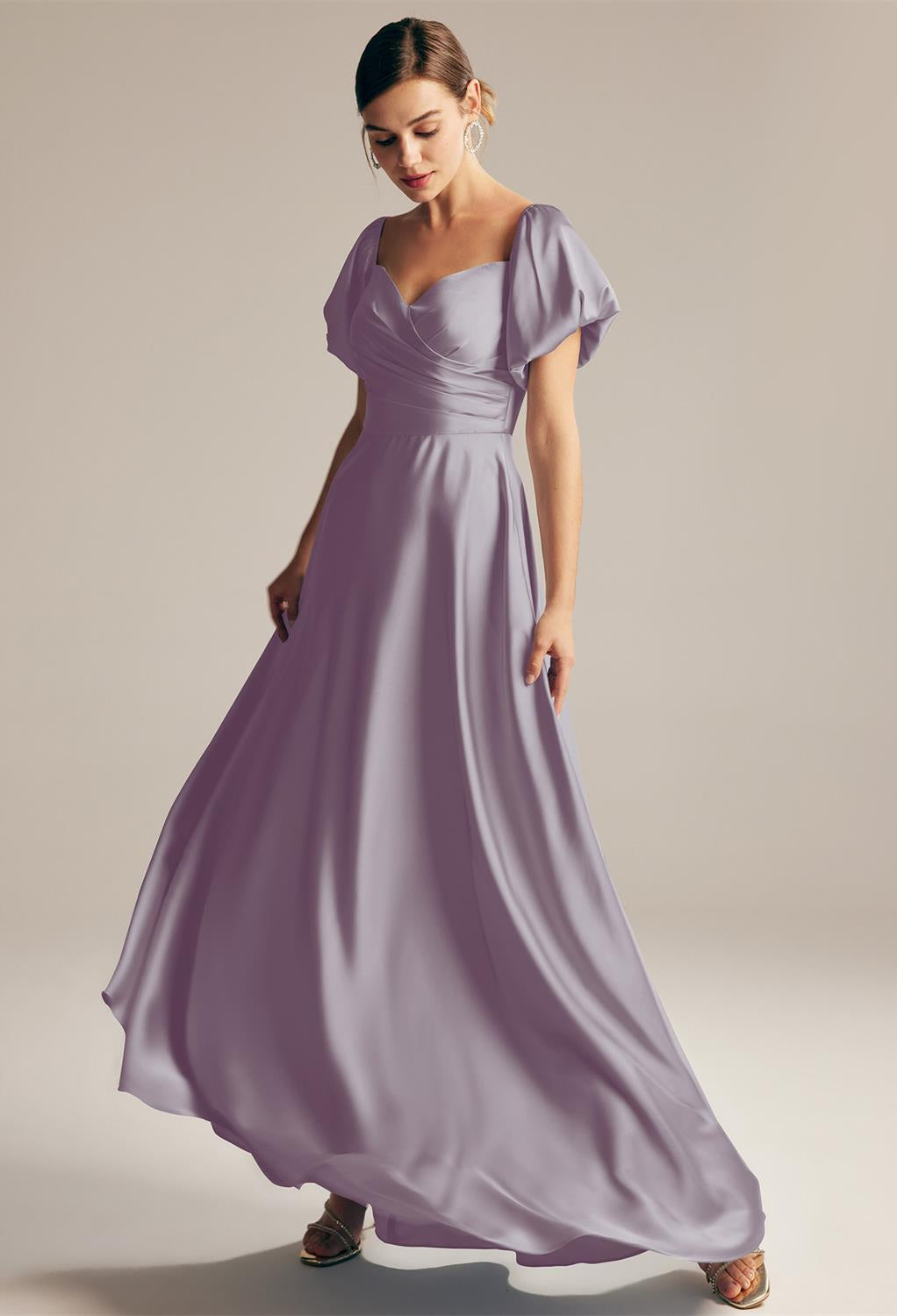 A woman in a flowing lavender dress from Bergamot Bridal's "Dey - Satin Charmeuse Bridesmaid Dress - Off The Rack" collection stands against a neutral background, looking down and slightly turned to the side.