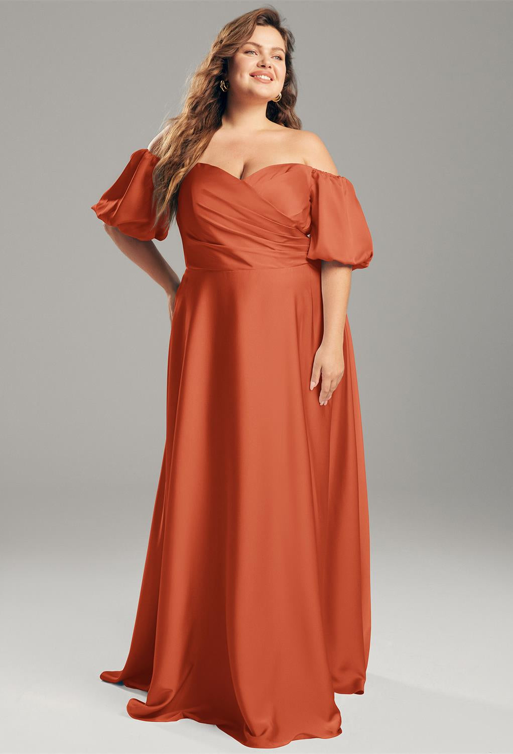 A person wearing the Dey - Satin Charmeuse Bridesmaid Dress by Bergamot Bridal stands against a neutral background, resembling an elegant off-the-shoulder gown from a chic bridal shop in London, Ontario.