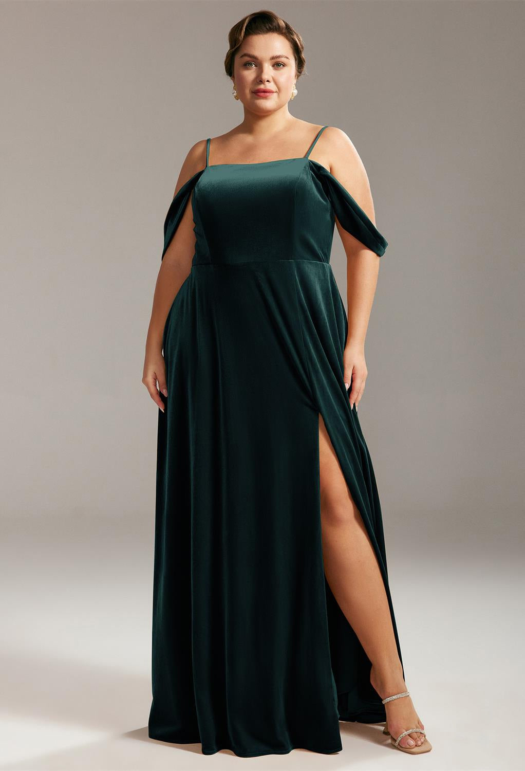 A woman in a Lowell - Velvet Bridesmaid Dress from Bergamot Bridal, with cold-shoulder sleeves and a thigh-high slit, standing confidently against a neutral background.