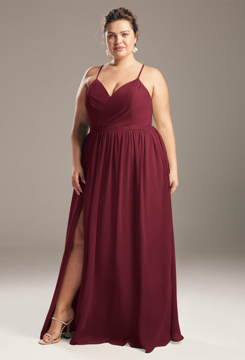 Decked out in Bergamot Bridal's Wilfreda Chiffon Bridesmaid Dress—off the rack—a person stands poised against a neutral background, exuding the elegance reminiscent of bridal shop finds in London, Ontario, with its sleeveless burgundy design and high slit.