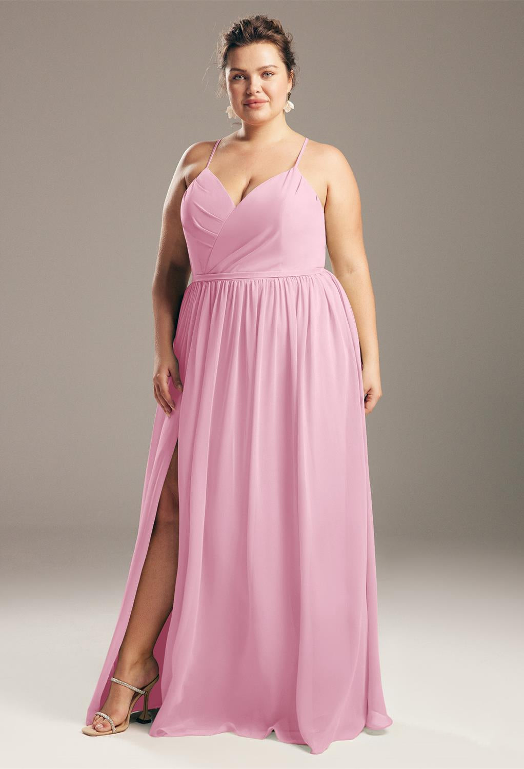 A woman stands in the Wilfreda Chiffon Bridesmaid Dress, an off-the-rack creation by Bergamot Bridal, featuring a pink sleeveless design with a side slit, reminiscent of the elegant styles found at bridal shops in London, Ontario.