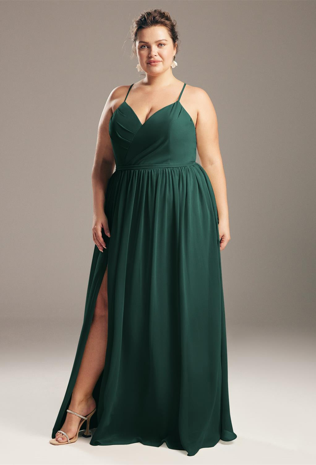 A woman in an elegant green wedding dress stands confidently, looking at the camera with one hand on her hip and a slight smile wearing the Bergamot Bridal Wilfreda Chiffon Bridesmaid Dress Off The Rack.