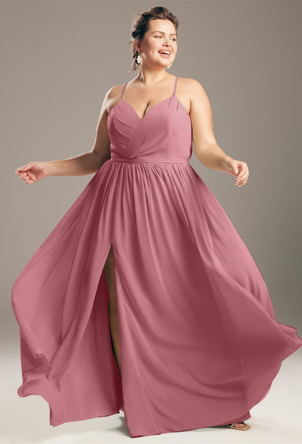 A woman in a flowing pink Wilfreda - Chiffon Bridesmaid Dress - Off The Rack from Bergamot Bridal in London smiling and posing playfully against a neutral background.